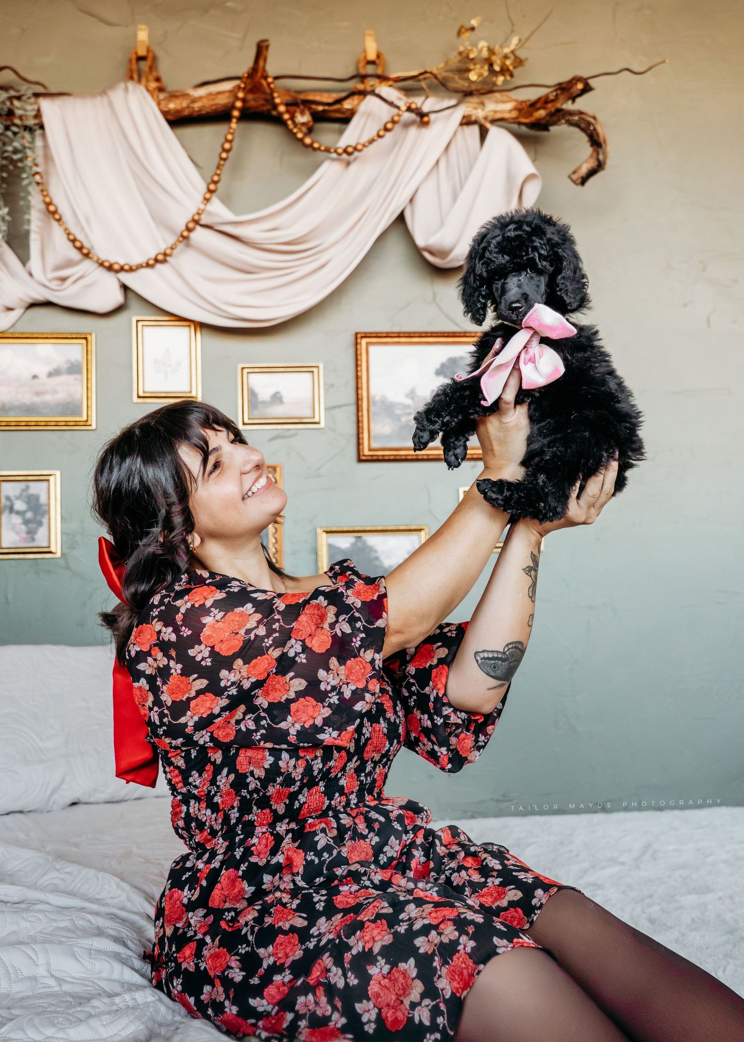 Person in floral dress playing with small black poodle puppy on a bed.