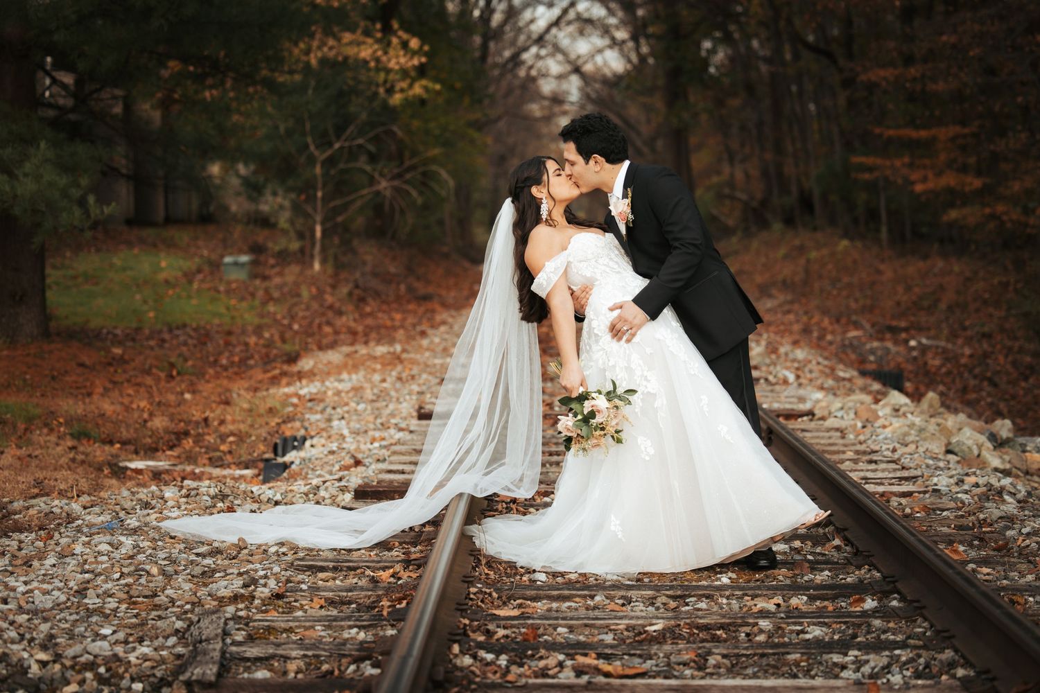 Romantic wedding couple poses on rustic railroad tracks surrounded by autumn foliage in scenic outdoor setting.