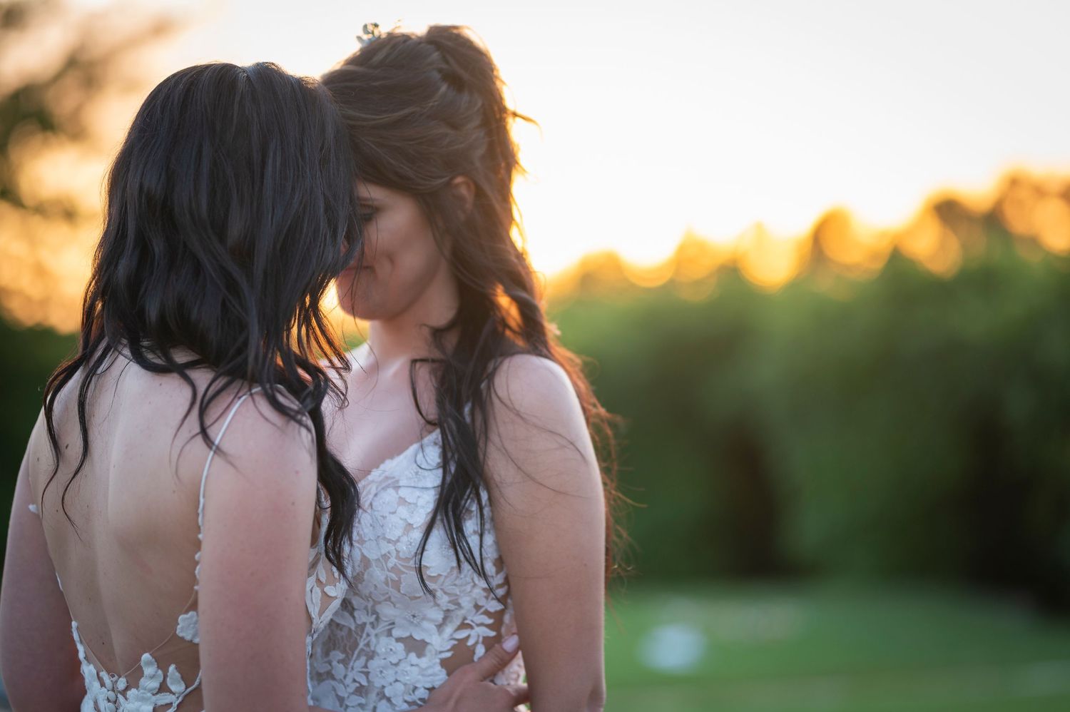 A couple in white wedding attire embraces during golden hour sunset in a grassy outdoor setting.