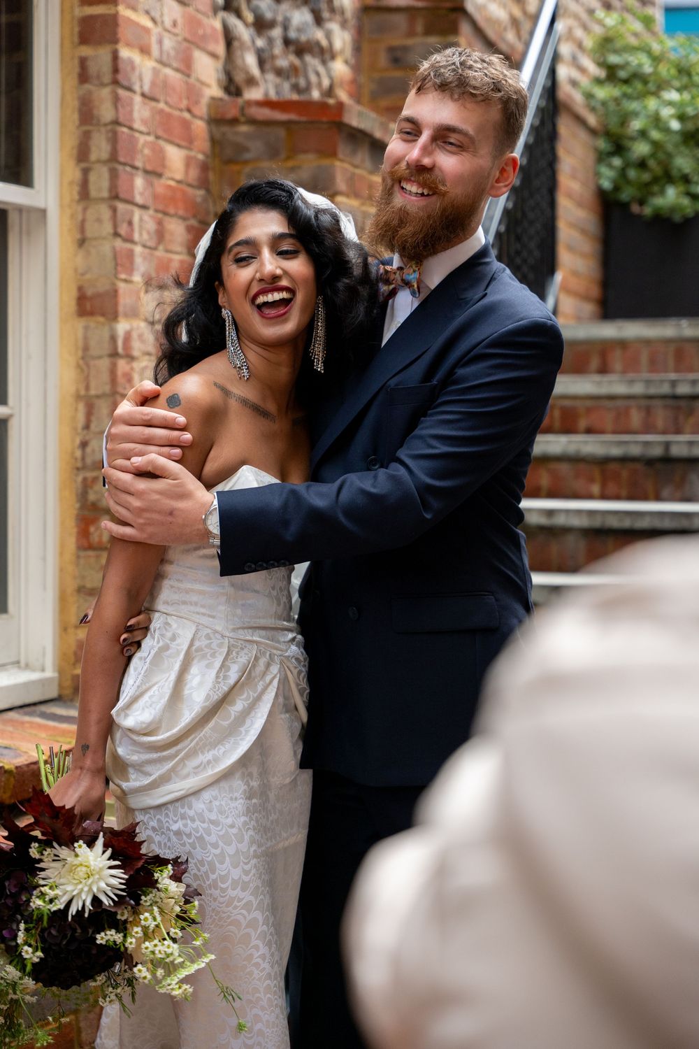 Joyful couple shares laughter during wedding celebration against brick wall backdrop.
