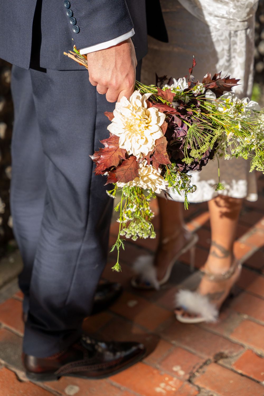 A person in a dark suit holds an elegant bouquet with peach dahlias and burgundy foliage against a brick floor.
