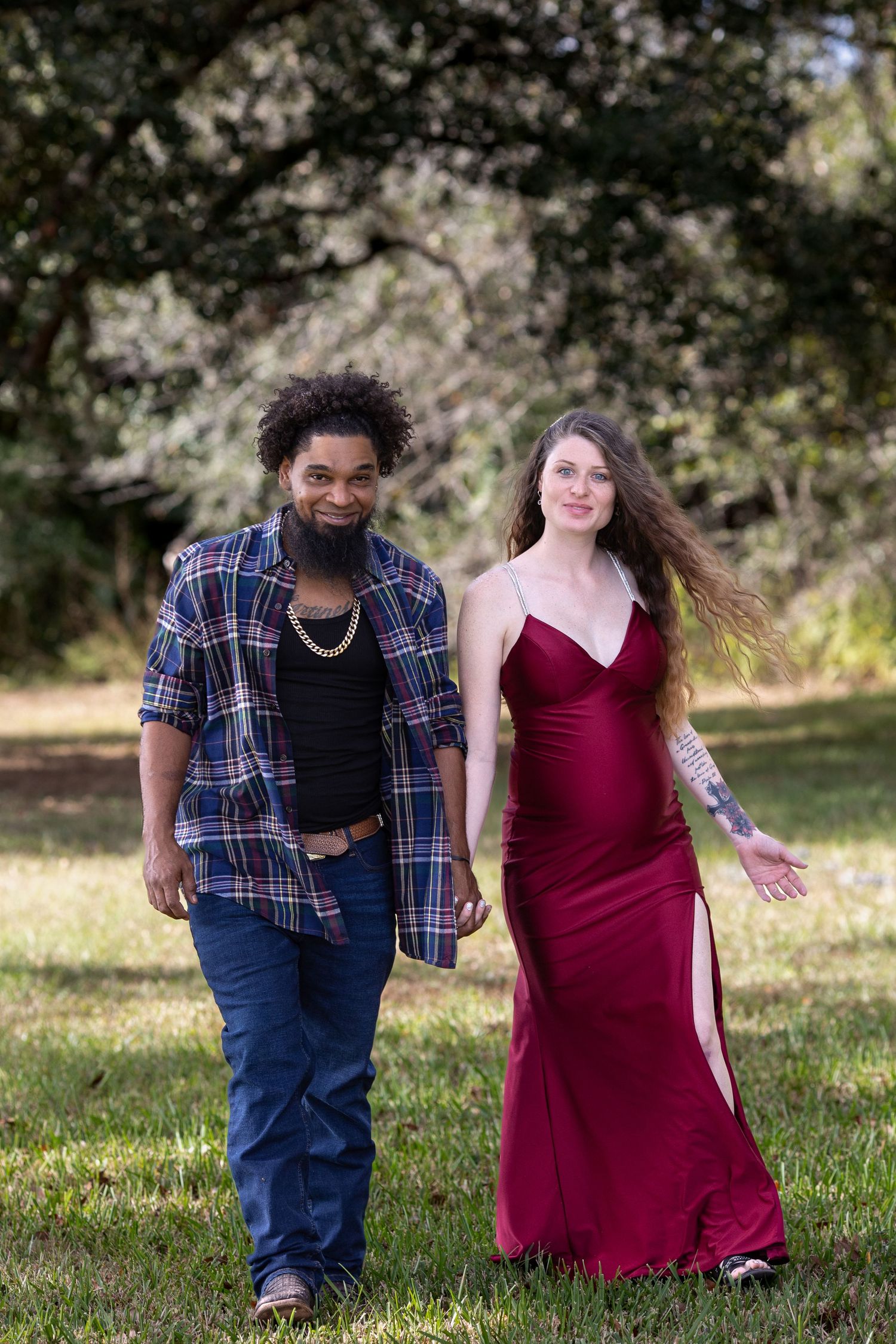 A couple in burgundy dress and plaid shirt sit together on grassy hill during outdoor photoshoot.