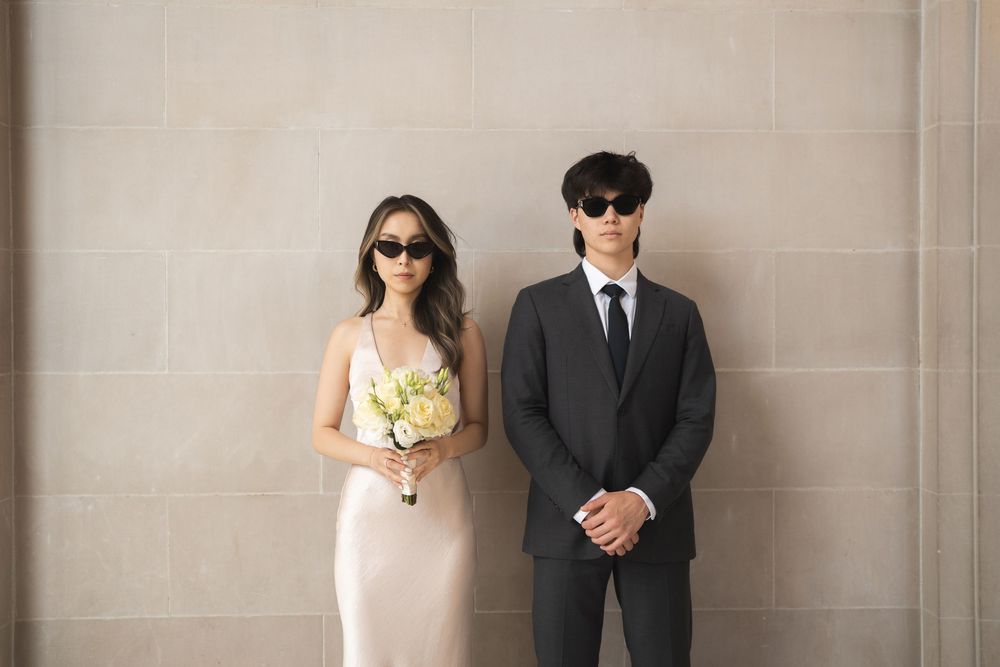 Bride and groom stand together in formal wedding attire against a neutral beige wall for elegant portraits.