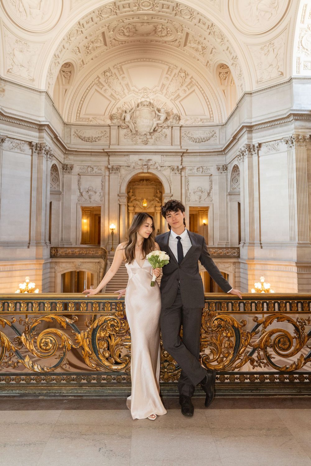 A couple poses in formal attire against an ornate baroque-style balcony with gilded details.