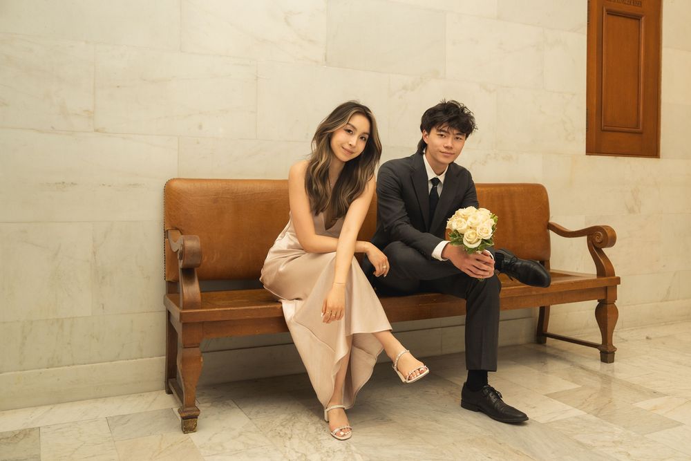 Couple sitting together on wooden bench in elegant formal attire at outdoor venue.