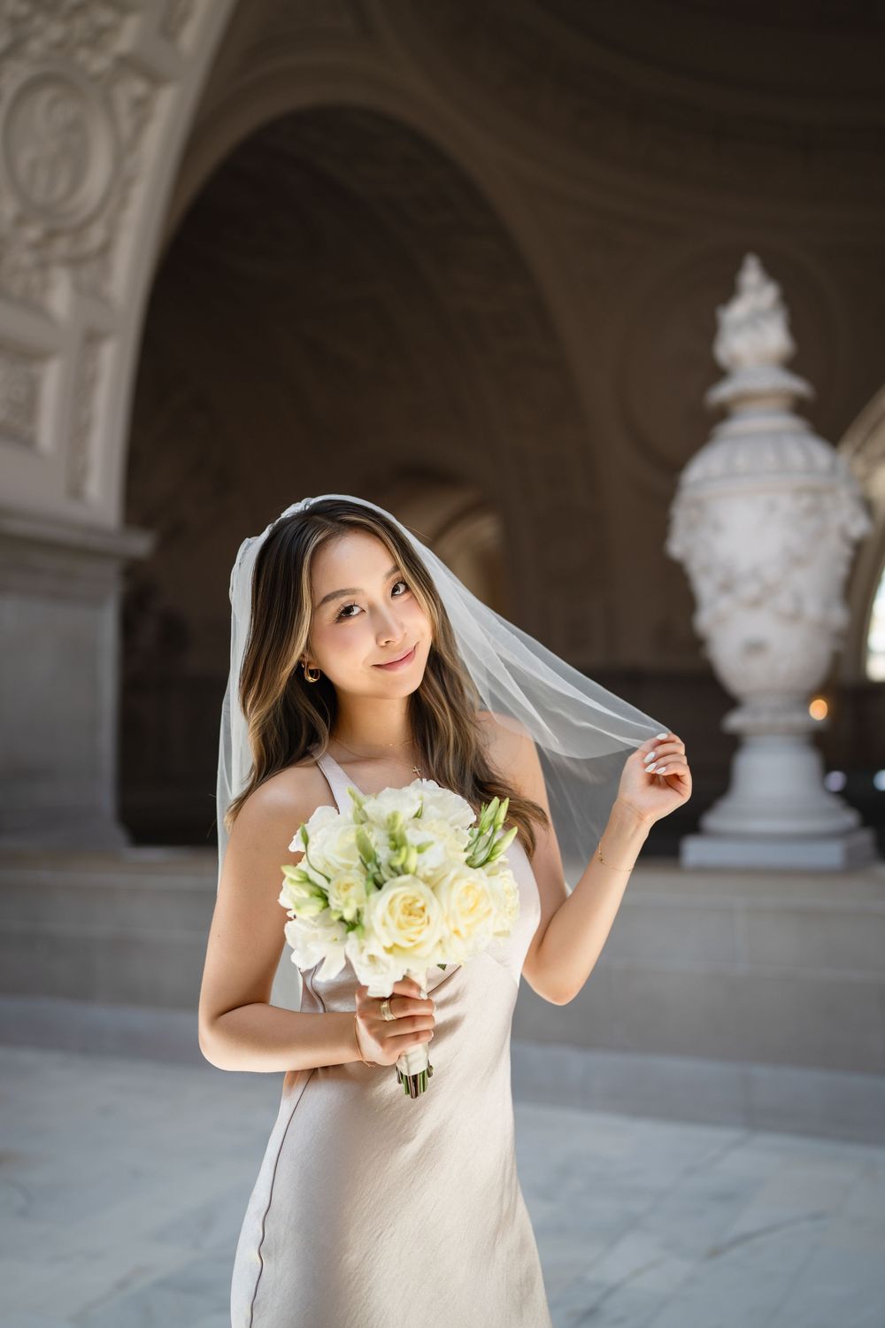 Bride in white dress holds cream-colored bouquet in marble-columned interior space.