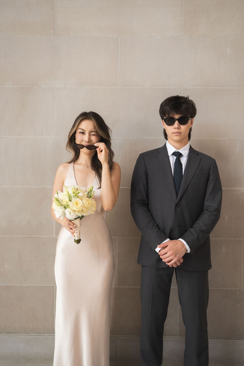 A couple poses for a wedding photo with the bride in white holding a bouquet while the groom wears sunglasses and a suit.