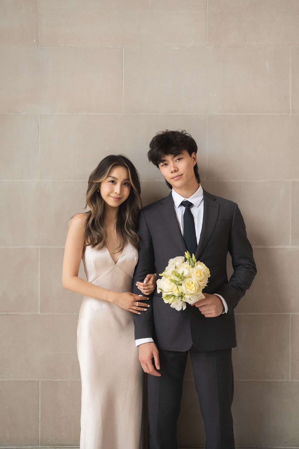 A couple in formal attire poses against a neutral wall while holding yellow bouquet flowers.