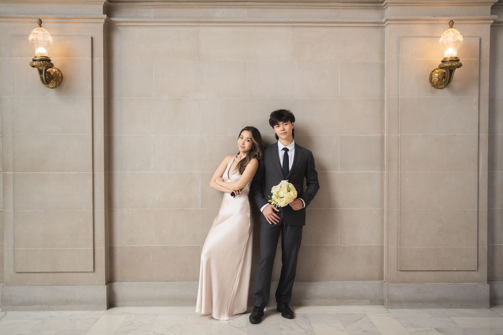 Elegant couple poses against a light gray wall with wall sconces in a formal setting.