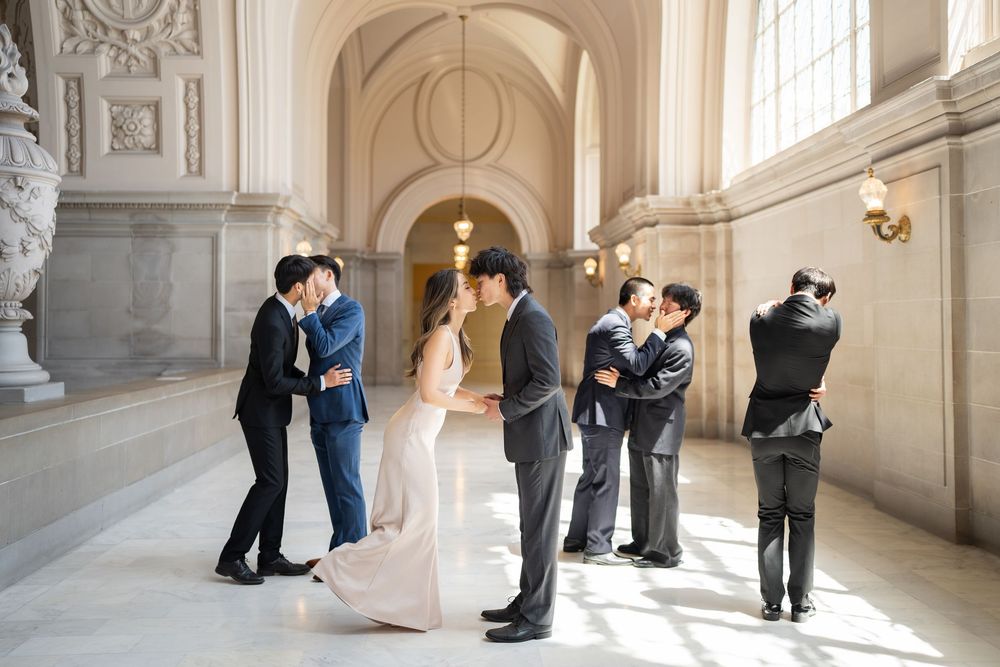 Elegant couples pose in a grand marble hallway with ornate architectural details and natural window light.