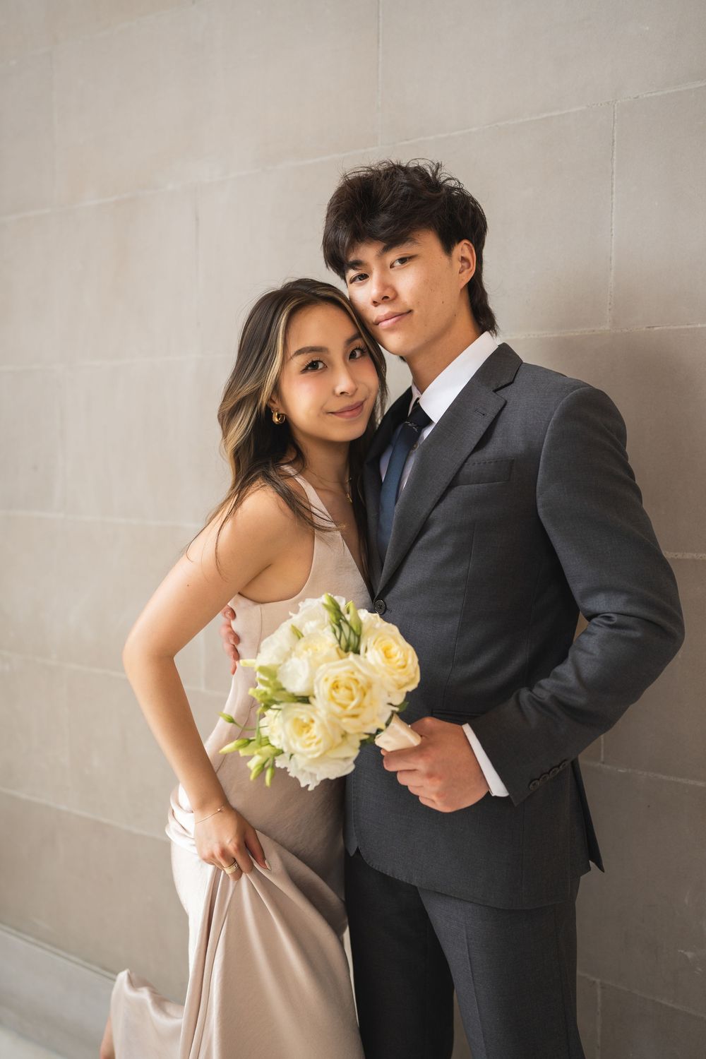 A romantic couple in formal wear poses together holding a bouquet of cream roses.