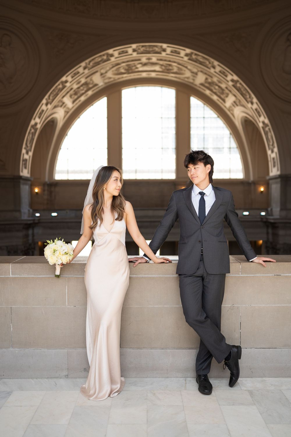 A couple in wedding attire sits and poses together on a stone ledge in an elegant architectural setting.