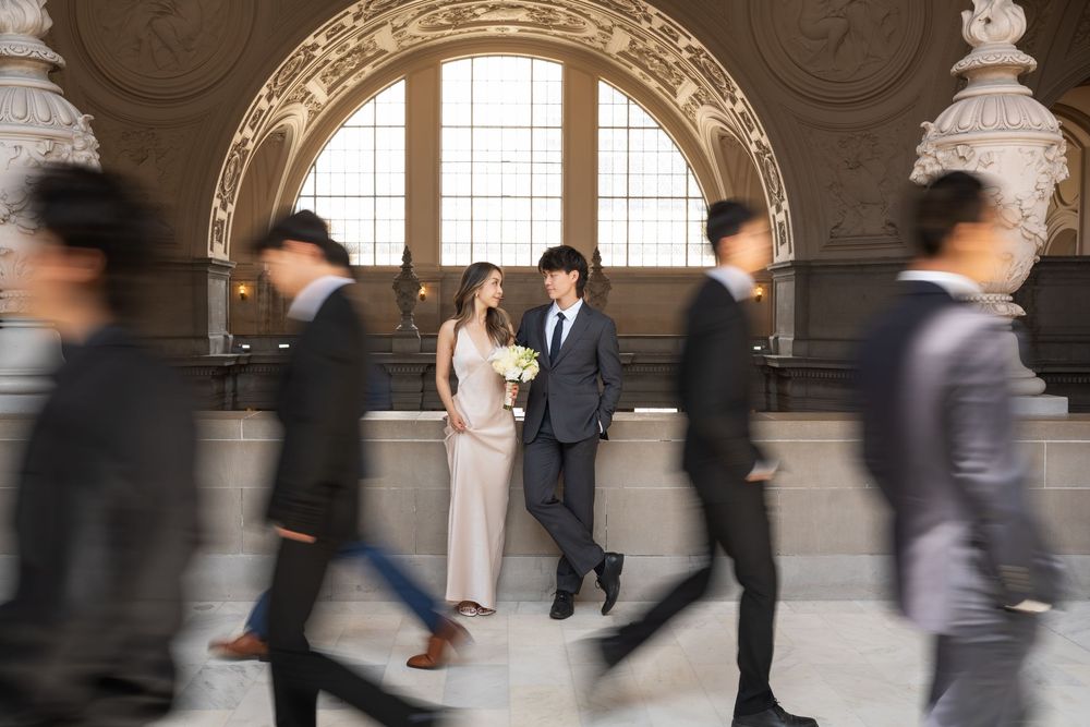 A couple poses in an elegant hallway while motion-blurred figures walk past them in formal attire.