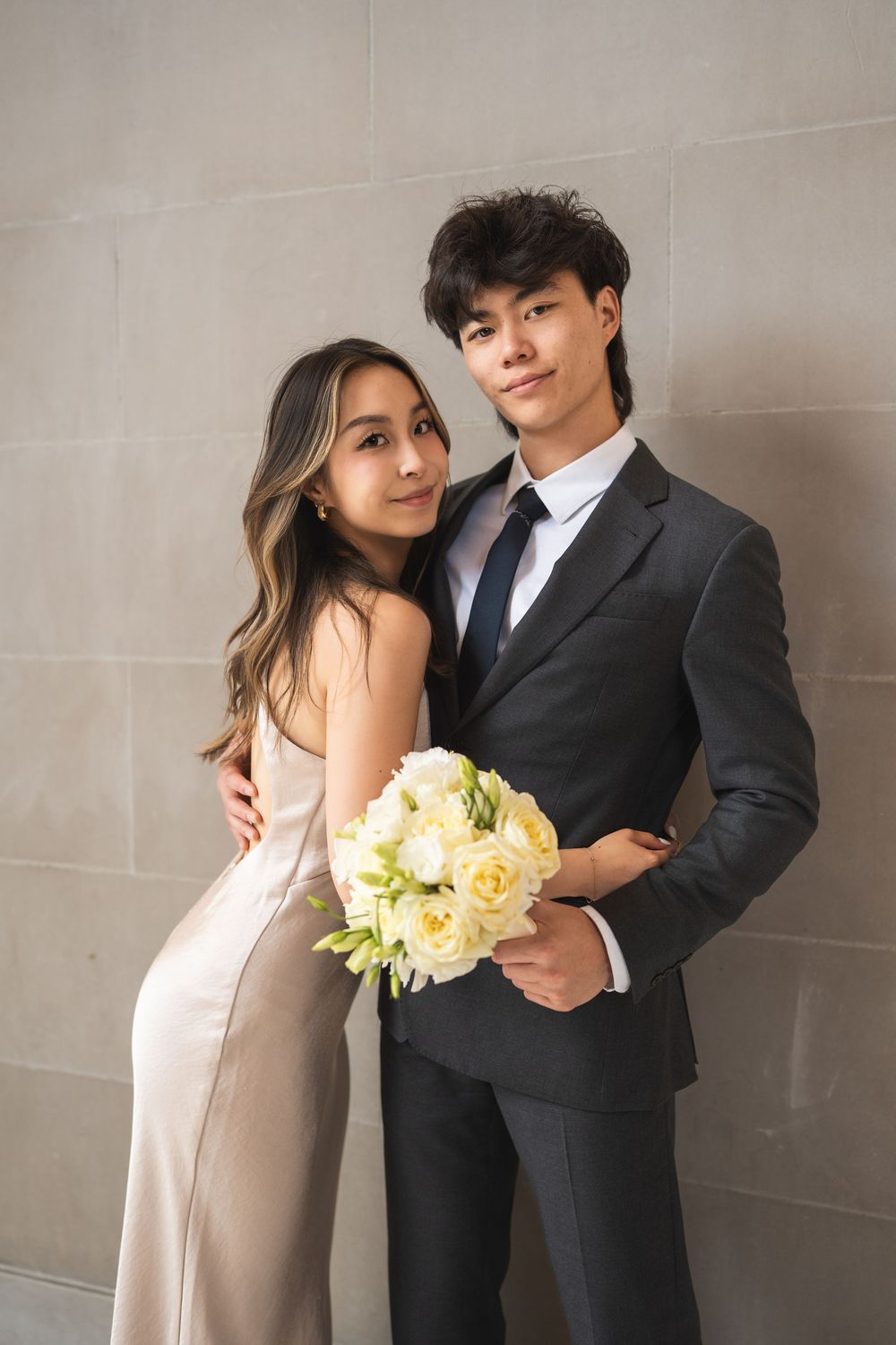 A couple in formal attire poses with a yellow and white bouquet against a stone wall.