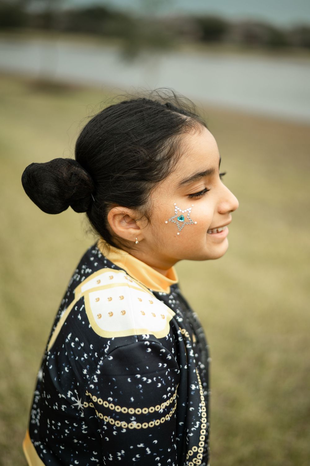 A side profile portrait showing sparkly face decorations and a black outfit with gold chain details against a blurred background.