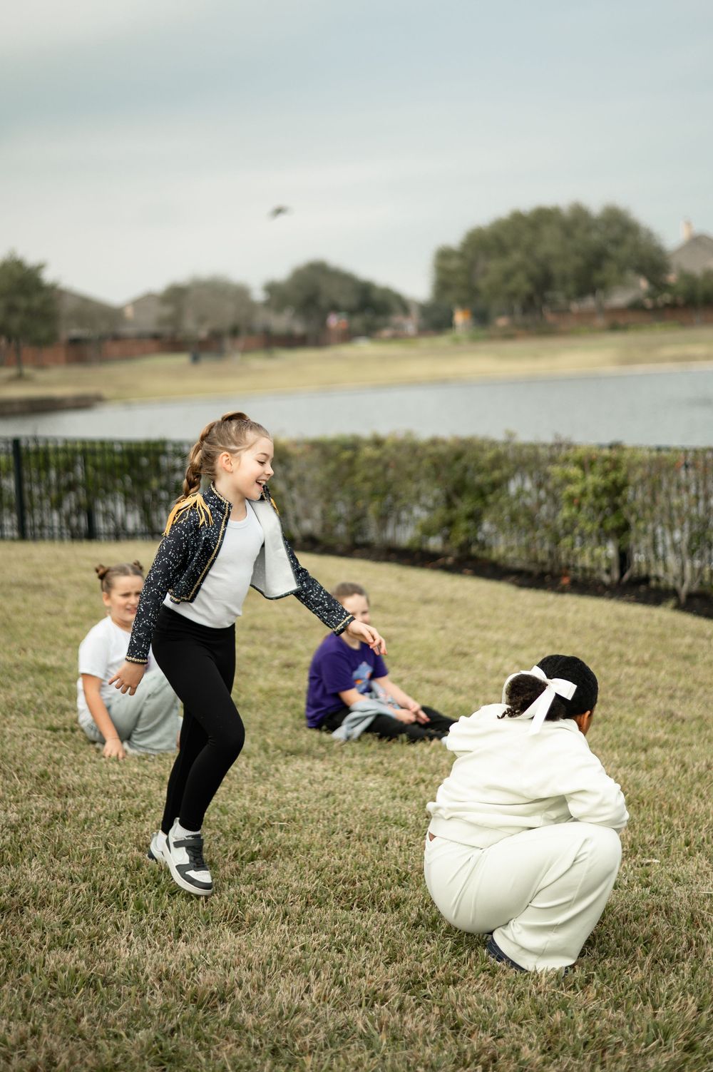 Children playing and running on a grassy field with a person in white sitting on the ground.