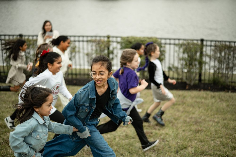 Children playing and running together on a grassy field with a fence in the background on an overcast day.