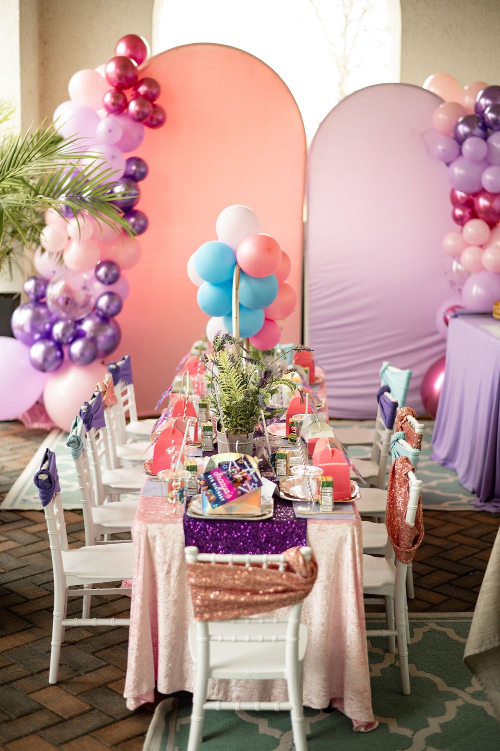 Elegant party table setup with pink and purple balloon arch, sequin tablecloth, and white chairs for a festive celebration.