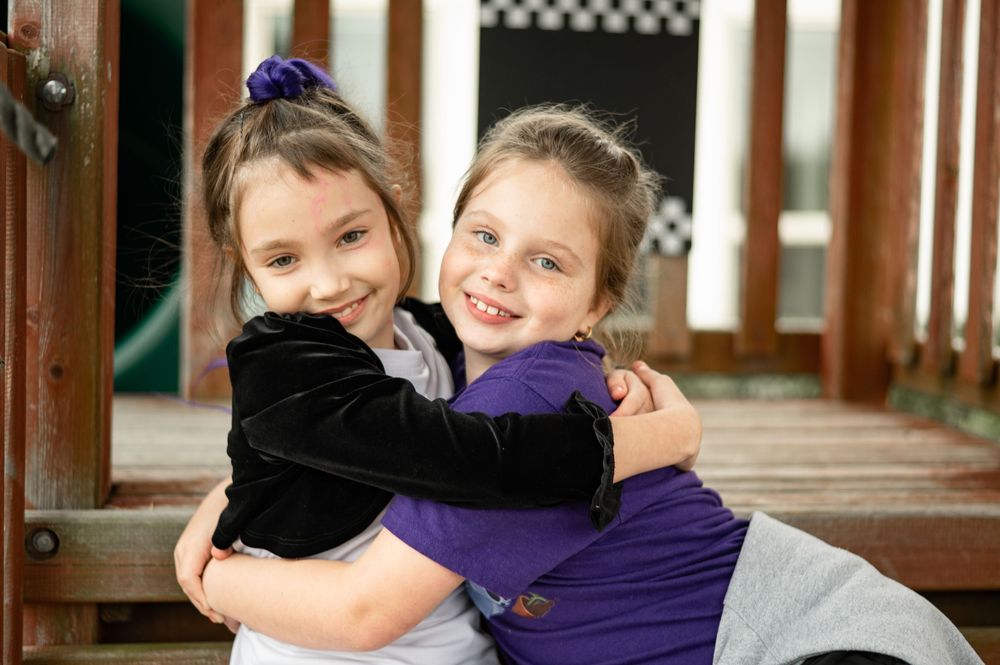 Two children in casual clothing share a warm embrace while smiling on a wooden porch.
