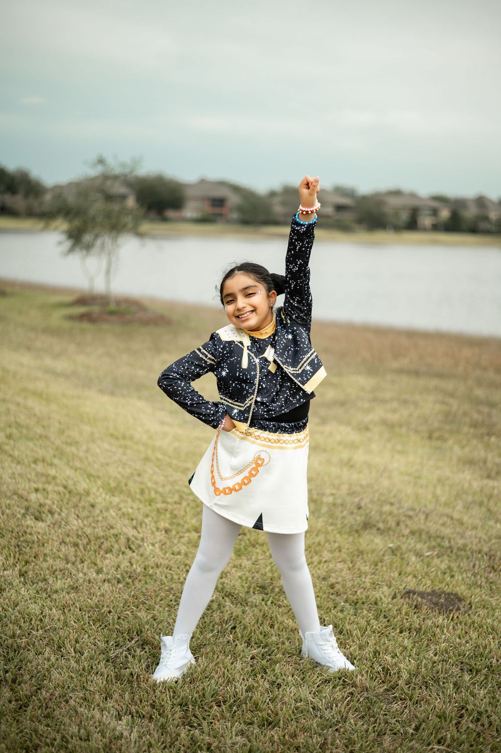 A child in a sparkly navy top and white pants strikes playful poses on grassy field near a lake.