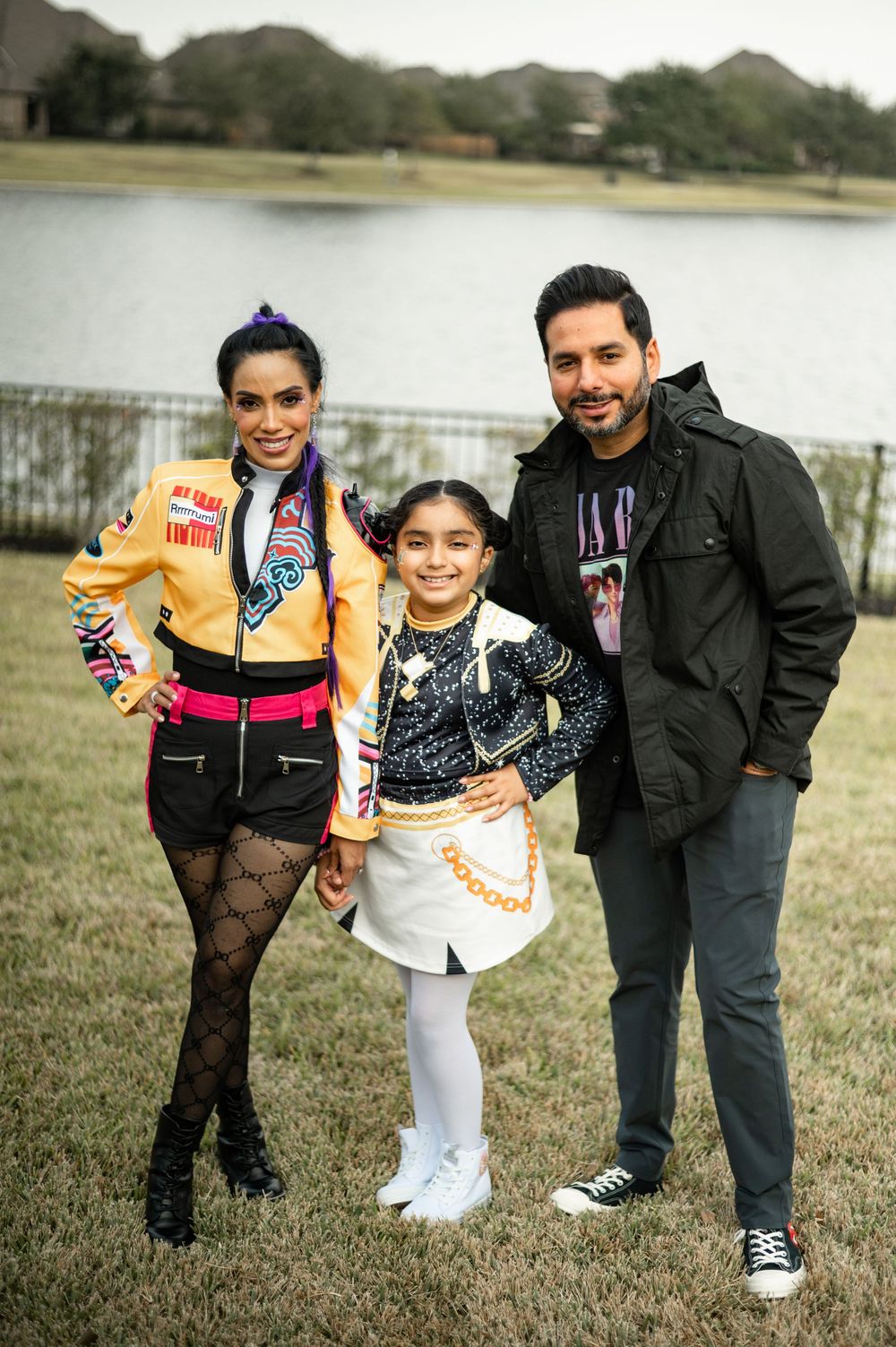 A family in casual winter attire poses together on grass near a lake, showing playful expressions and movements.