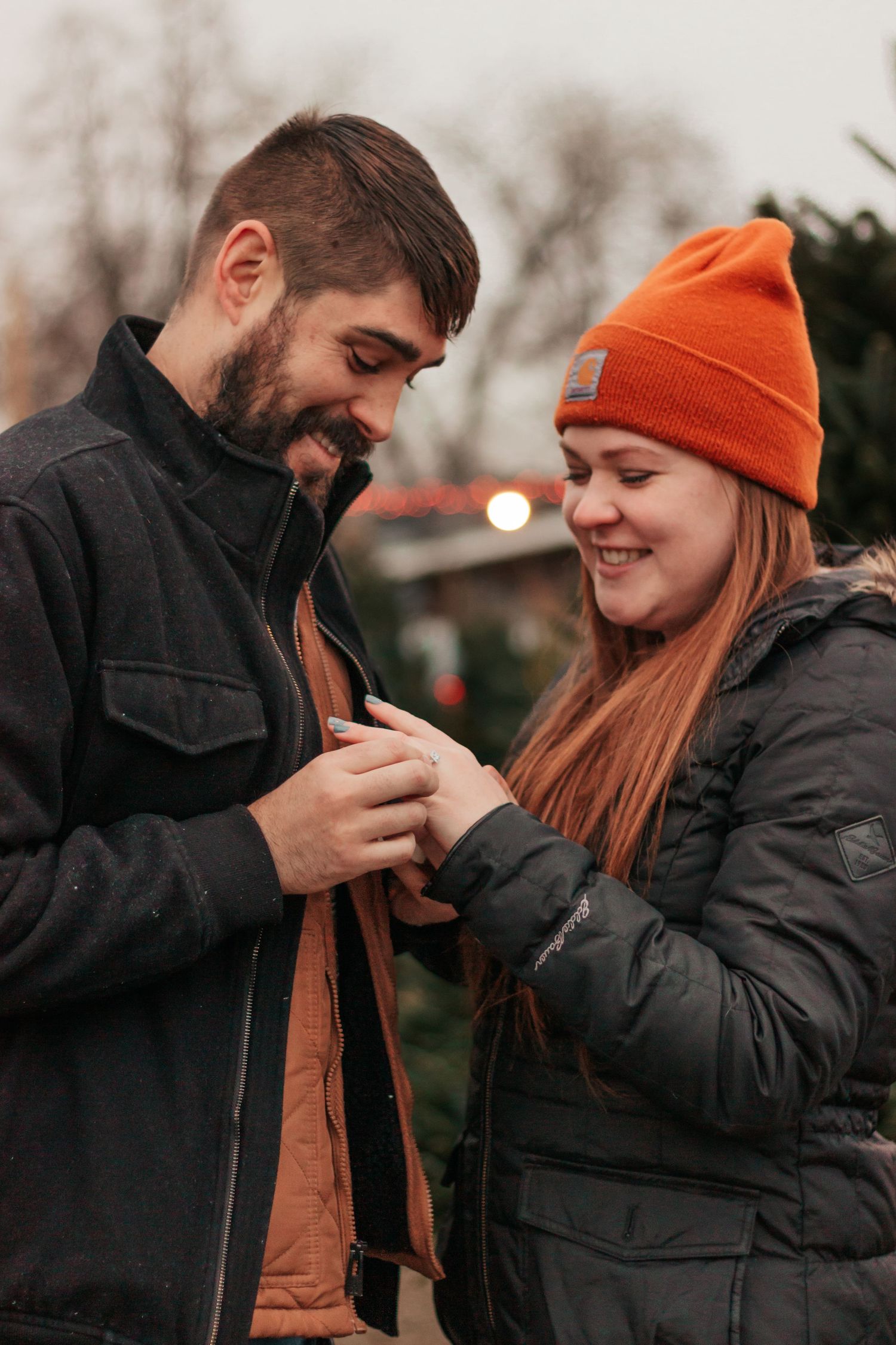 smiling couple in winter jackets examines an engagement ring outdoors near a Christmas tree lot| Westminster Colorado Proposal Photographer