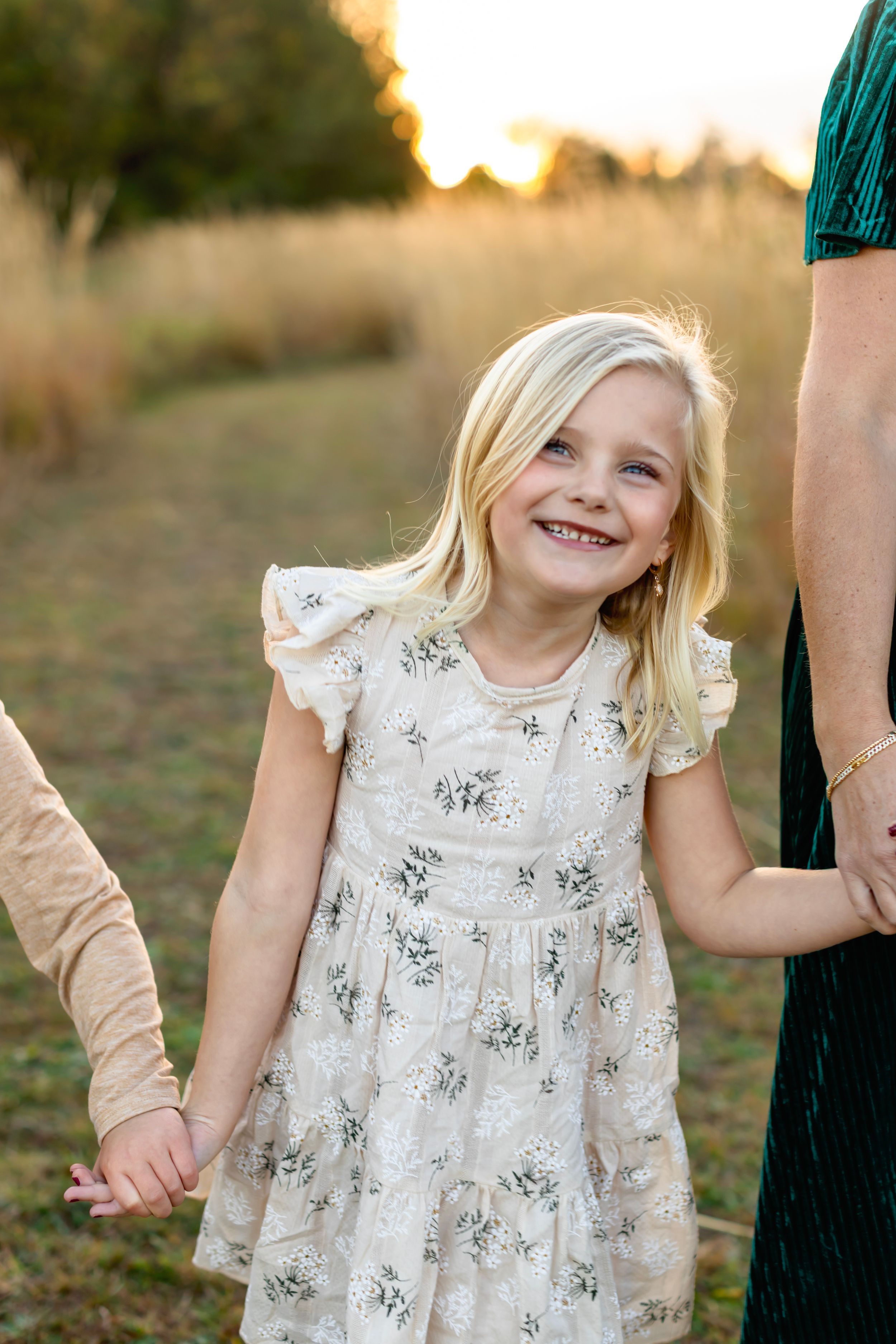 A joyful child in a floral dress holds hands with family members at sunset in a field.