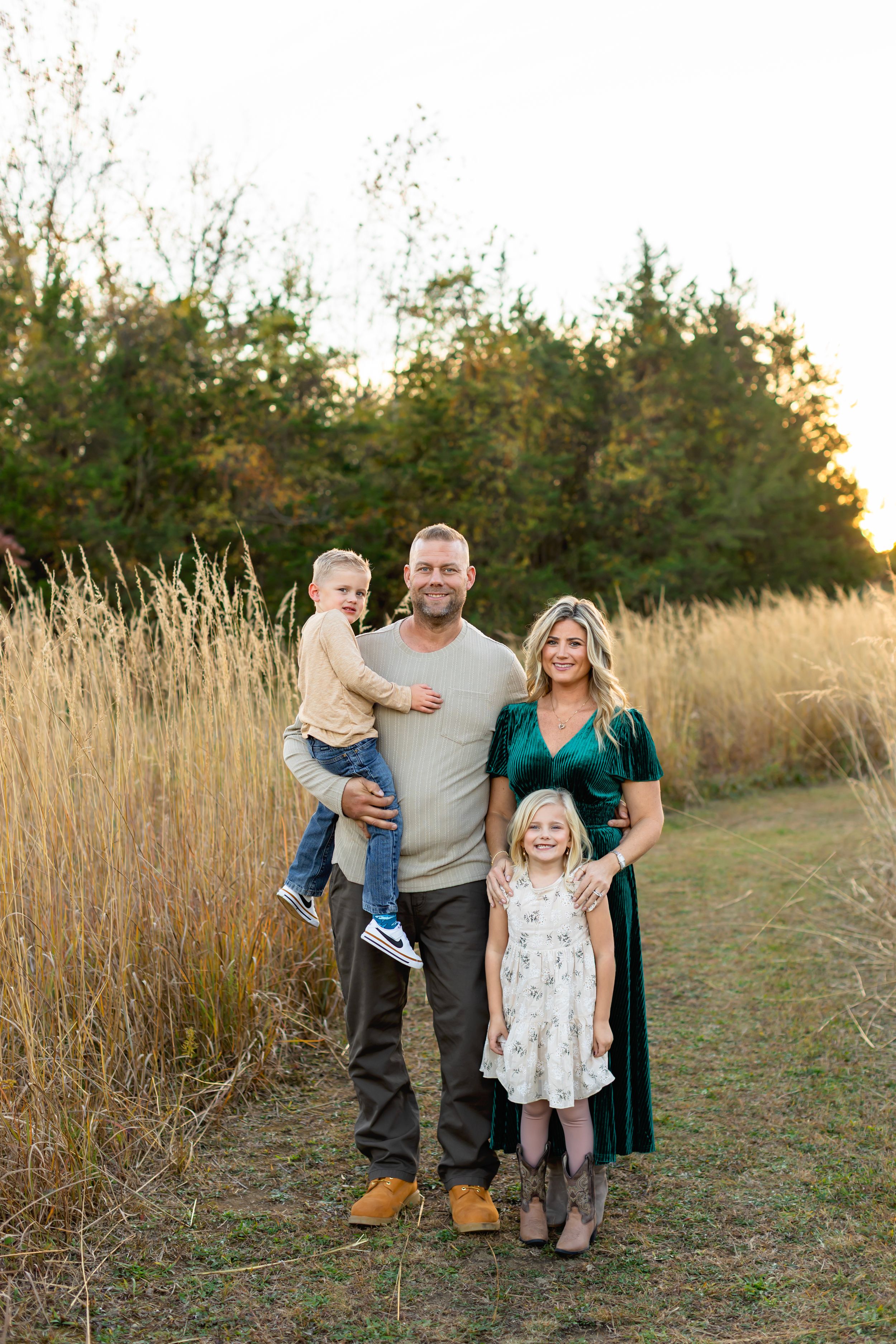 Family poses together in a grassy field with tall golden grass and trees at sunset.