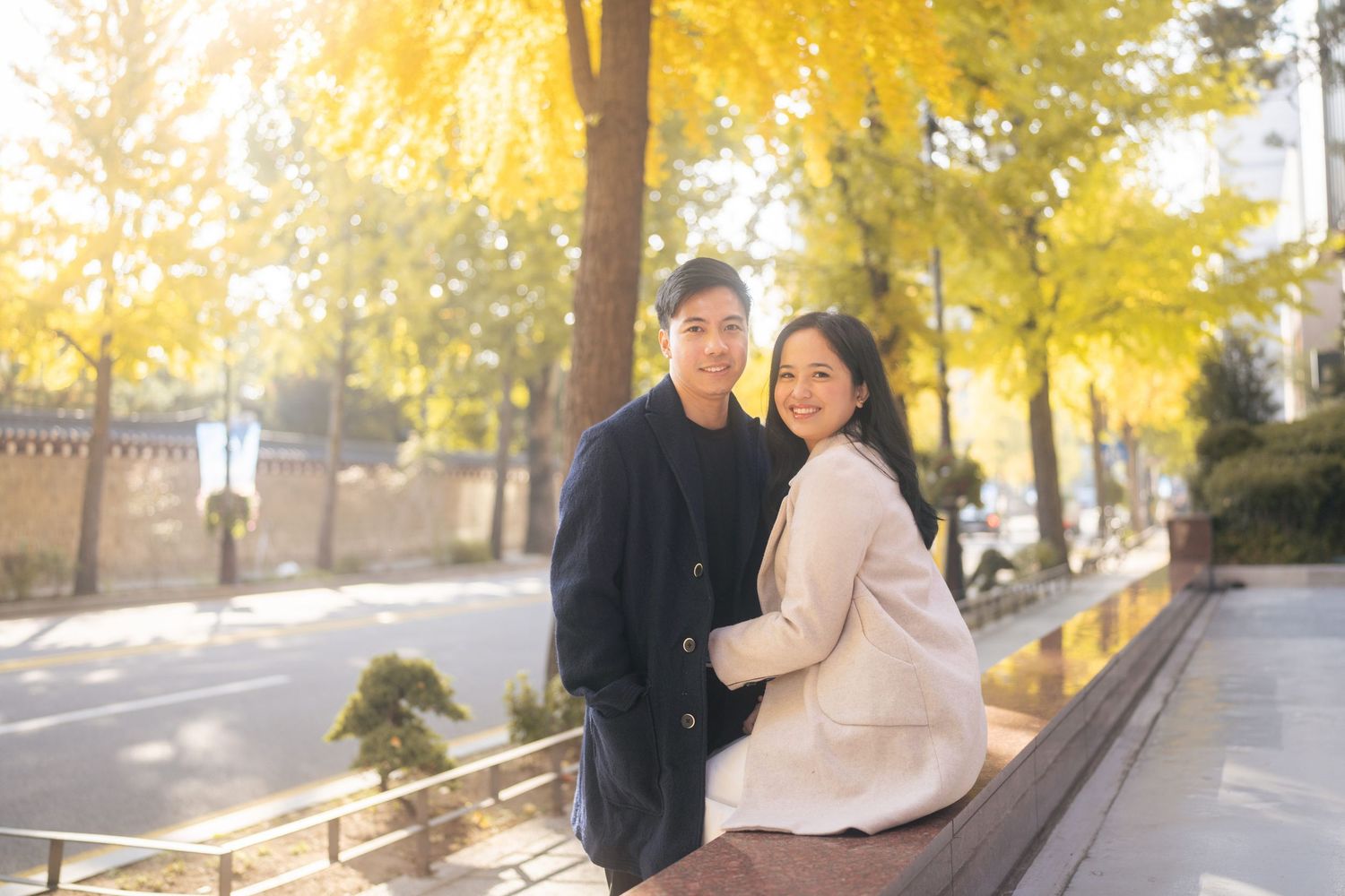 A couple embraces on a walkway surrounded by golden autumn trees and dappled sunlight.