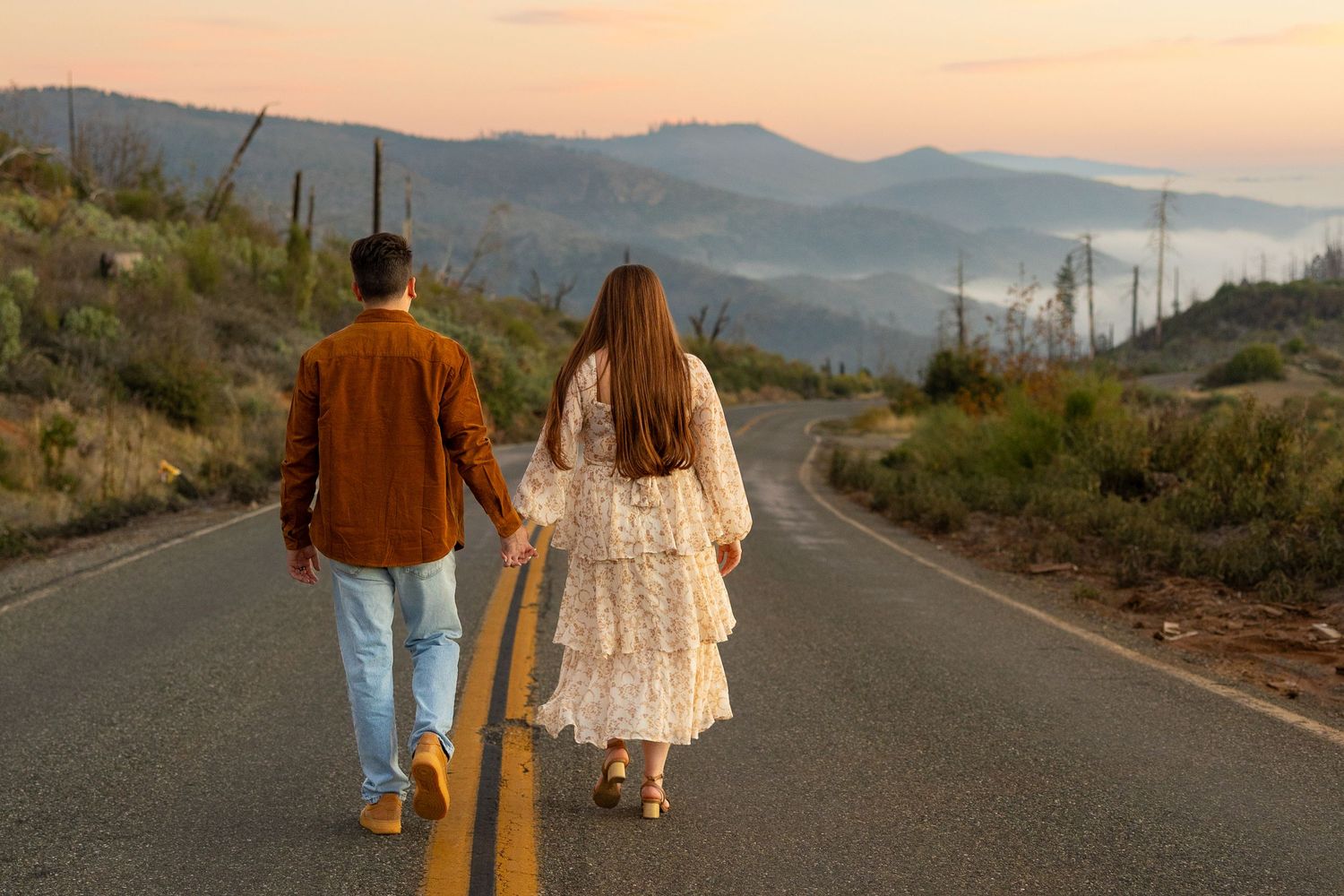 A couple walks hand in hand down a desert road at sunset with mountains in the distance.