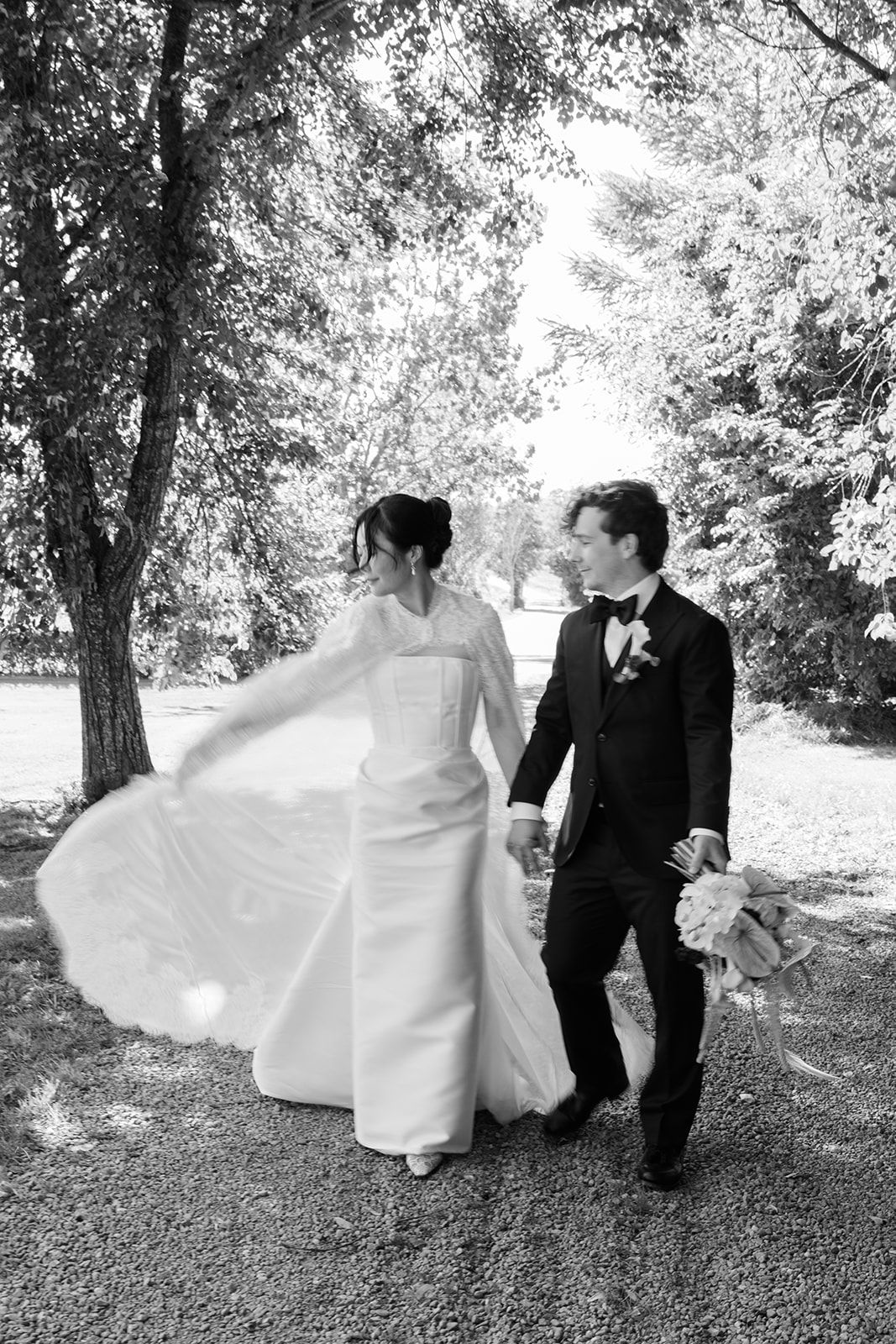 A black and white wedding photo showing a couple walking together outdoors with the bride's dress flowing in the breeze.
