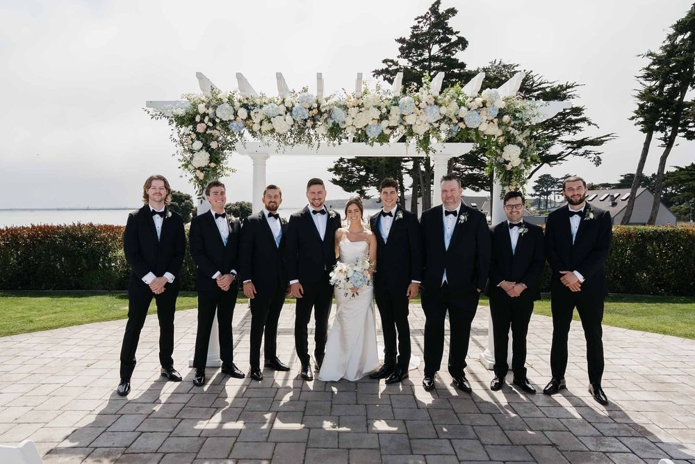 Wedding party poses under a white floral pergola on a stone patio during an elegant outdoor ceremony.