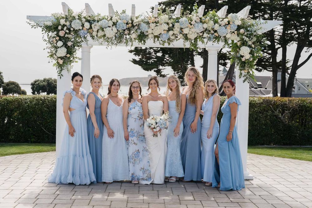 Wedding party wearing light blue bridesmaid dresses pose together outdoors on a sunny day.