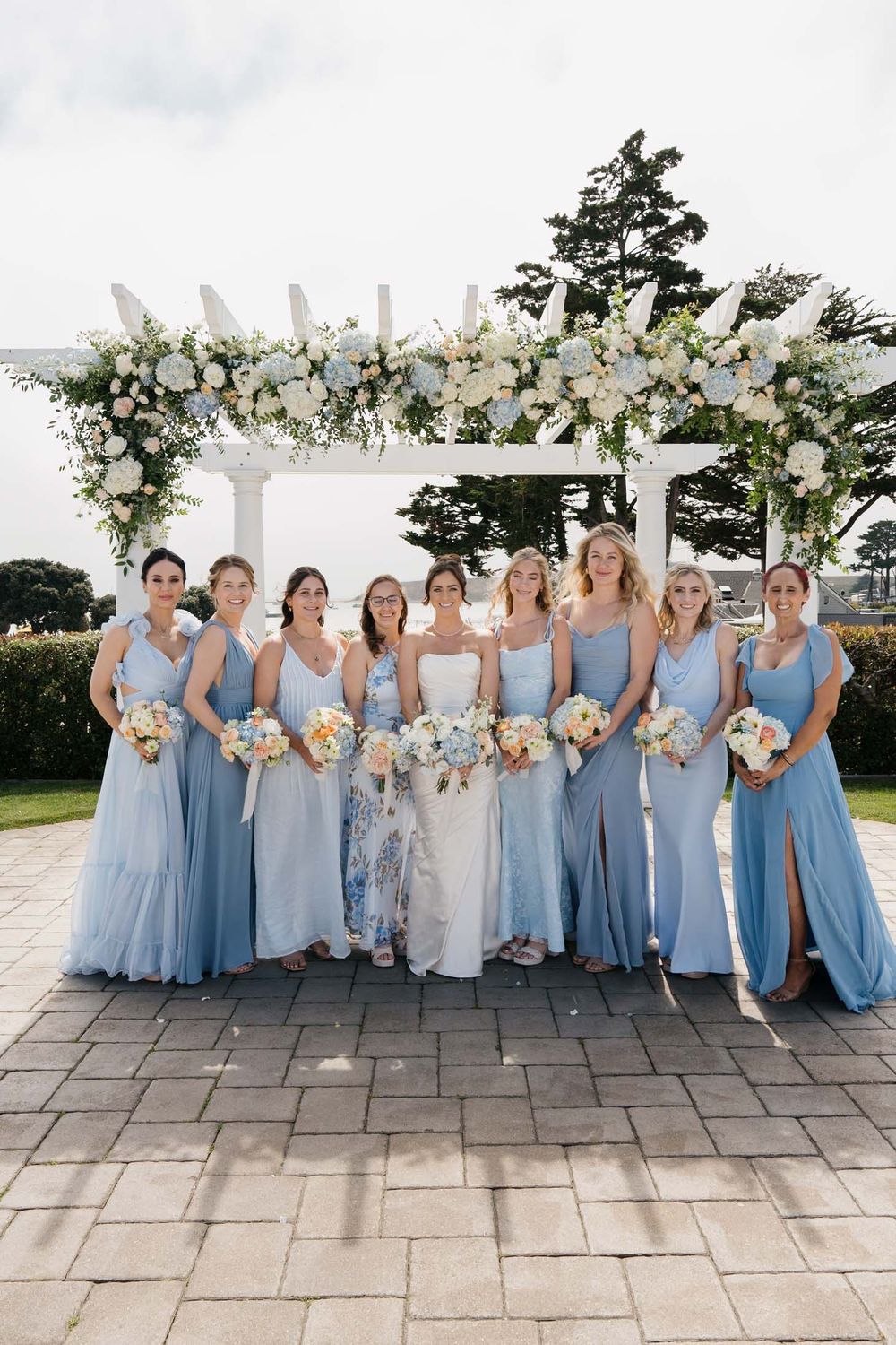 Wedding party wearing light blue bridesmaid dresses poses together on stone patio during outdoor celebration.
