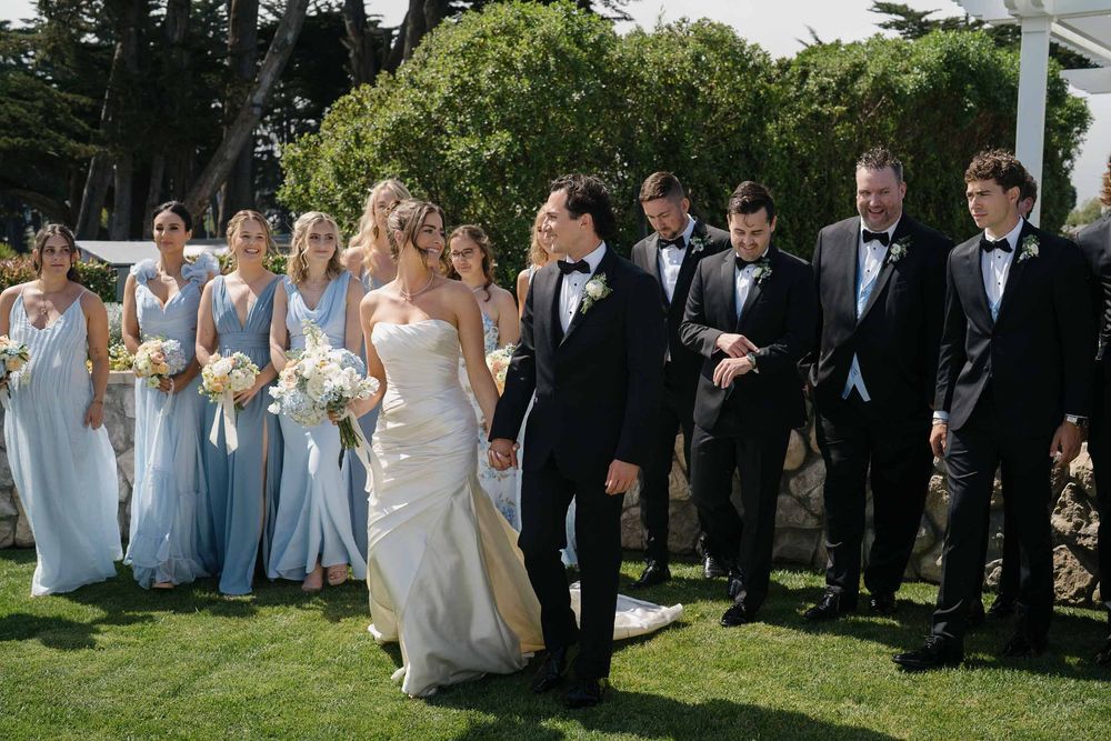 Wedding party members gather outdoors on a grassy lawn for formal group photos on a sunny day.