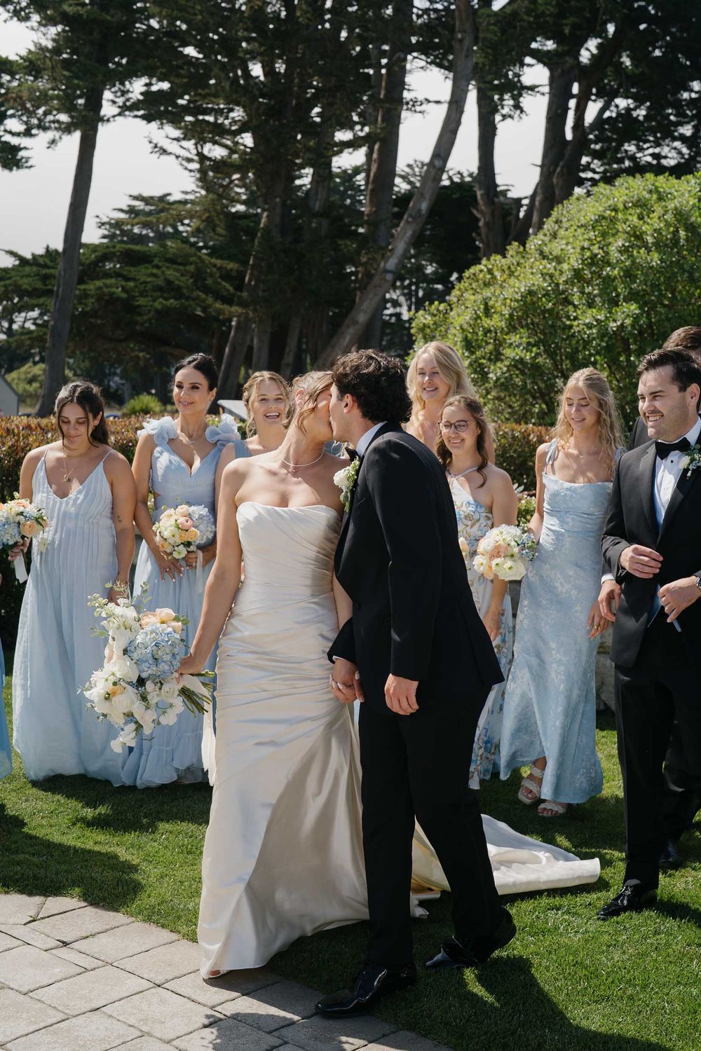 Wedding party in light blue bridesmaid dresses and black tuxedos poses on a sunny lawn with trees in background.
