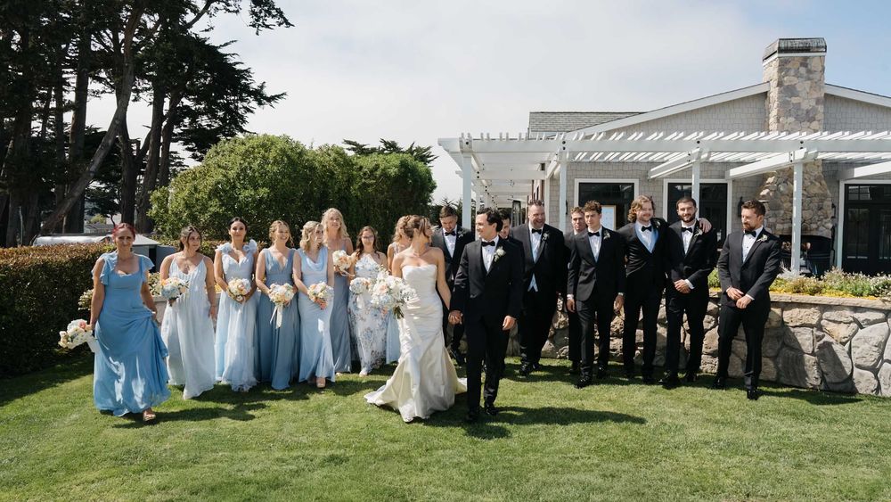 Wedding party in light blue dresses and black tuxedos gather on manicured lawn near coastal venue.
