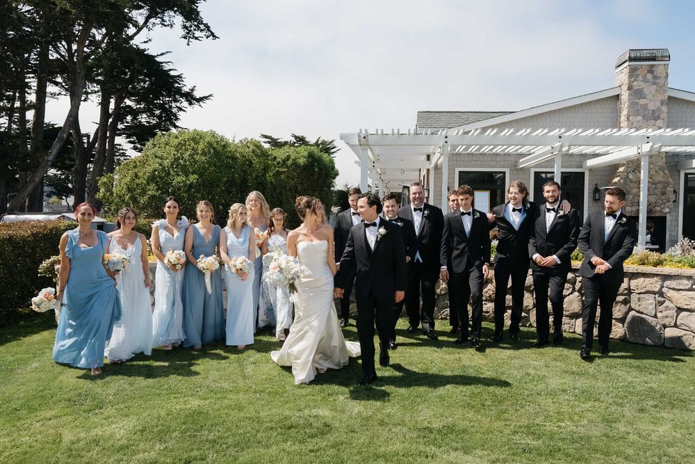 Wedding party members line up on a grassy lawn for formal group photos at an outdoor venue with white pergola.