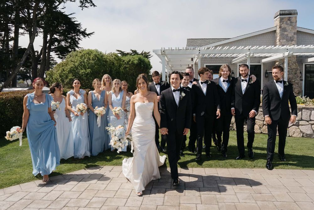 Wedding party members in light blue dresses and black tuxedos pose together outdoors at a coastal venue.