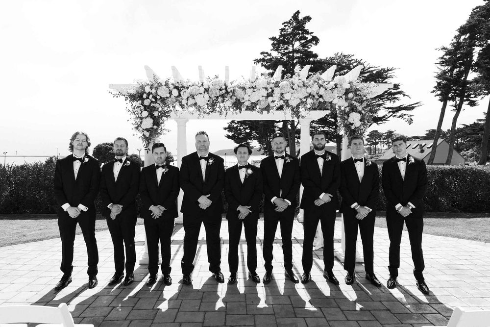 Black and white panoramic photo showing a wedding party lineup outdoors with trees in the background.