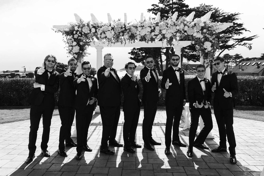 Black and white panoramic wedding photo showing formal group posing outdoors with trees in the background.
