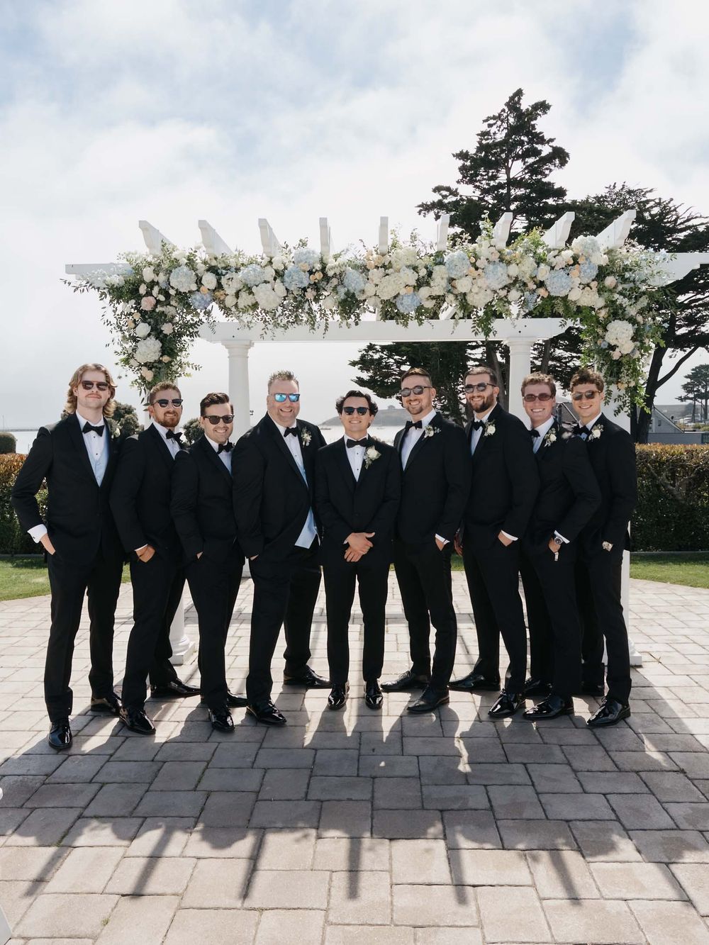 A wedding party in black tuxedos stands in front of a white floral arch on a stone patio during an outdoor ceremony.