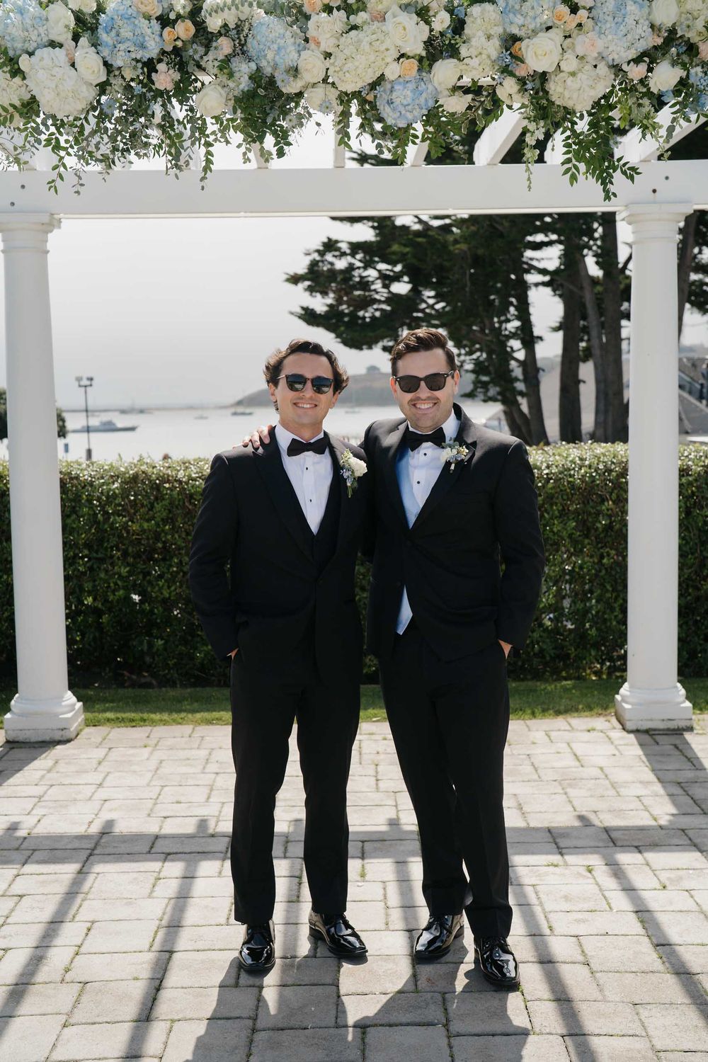 Wedding party members in formal attire stand under a white pergola with floral decorations.