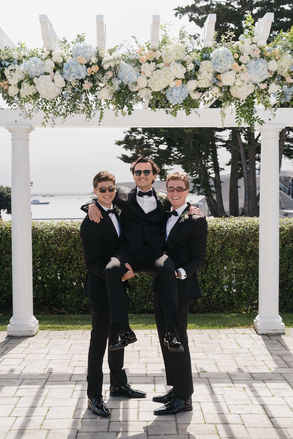 Groomsmen in black tuxedos and sunglasses pose casually under a white pergola by the ocean.