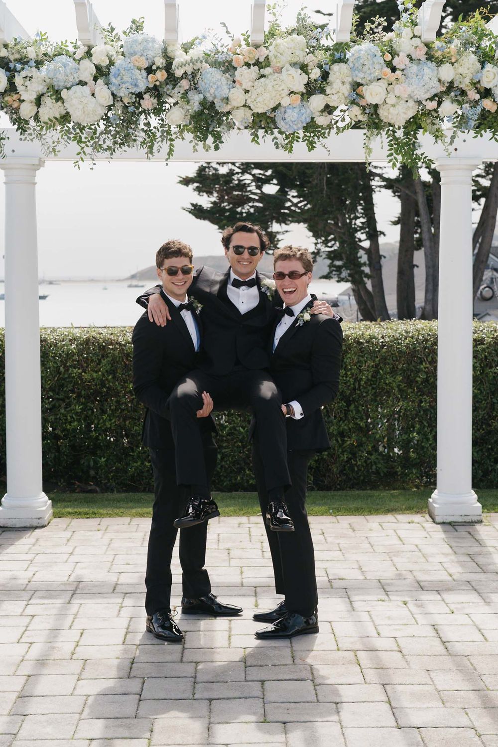 Groomsmen in black suits and sunglasses pose together under a white floral arch at an outdoor wedding venue.