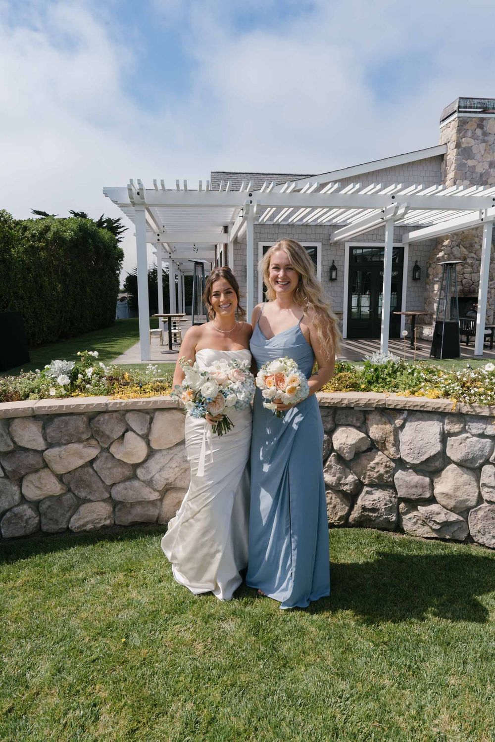 Two people in wedding attire pose together on a grassy lawn with a stone wall and white pergola in the background.