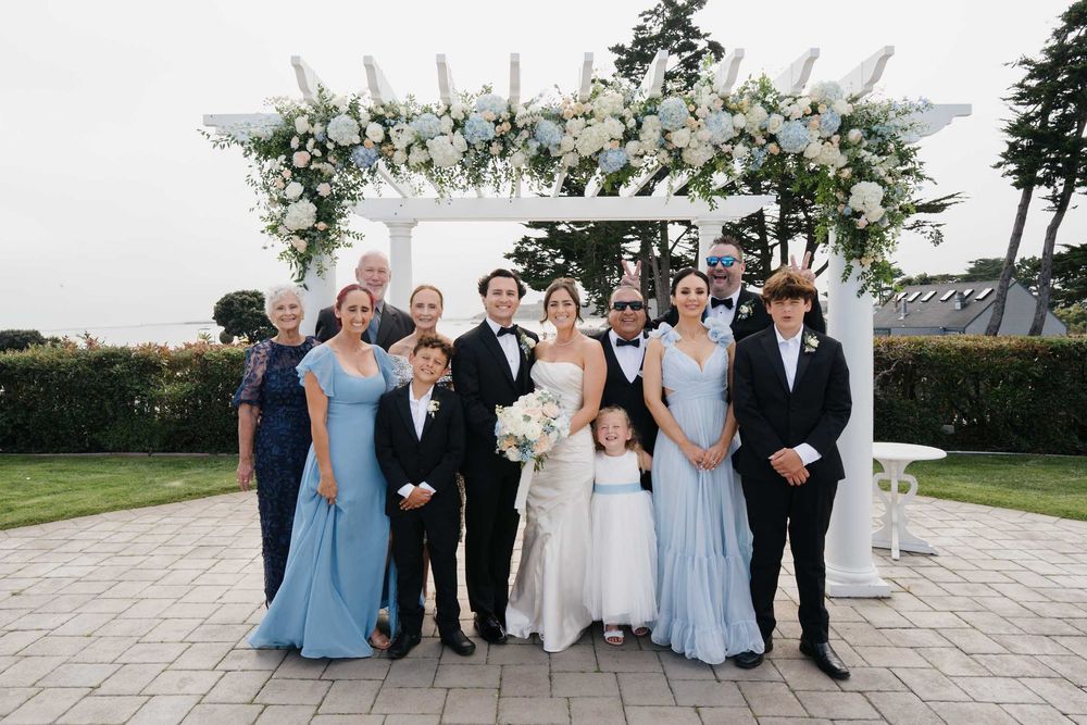 Wedding party members in blue dresses and black suits pose together outdoors for a formal group photo.
