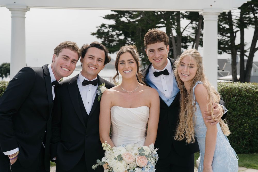 A group in formal wedding attire poses together on a white columned porch with trees visible in the background.