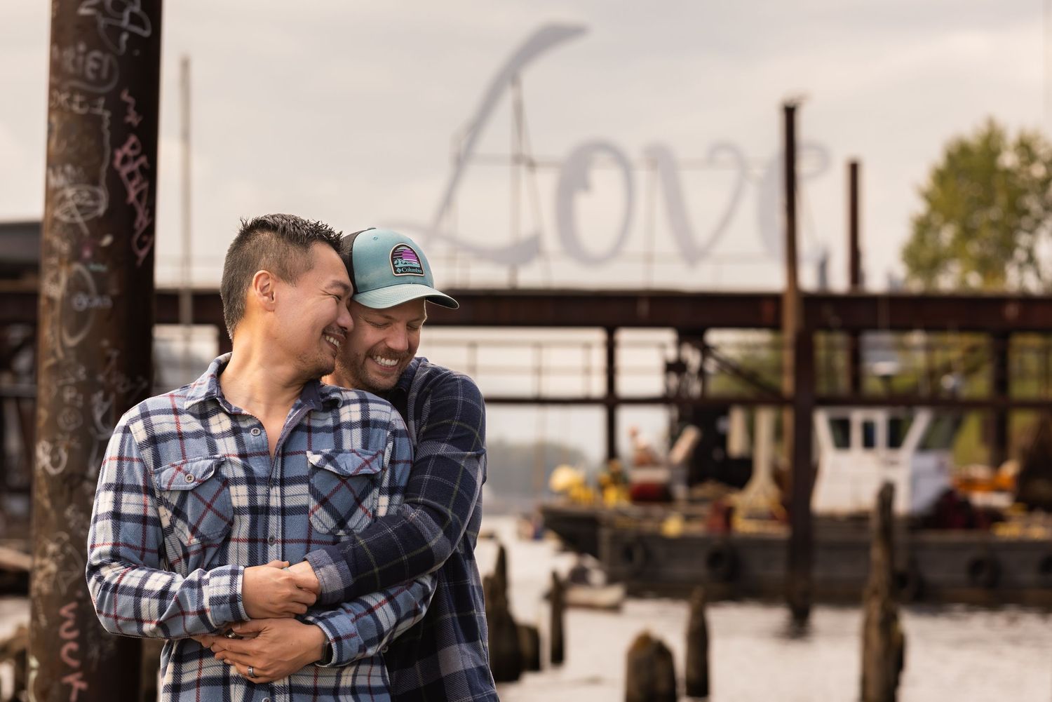 A couple in plaid shirts embraces while standing on a wooden dock near a lake at sunset.