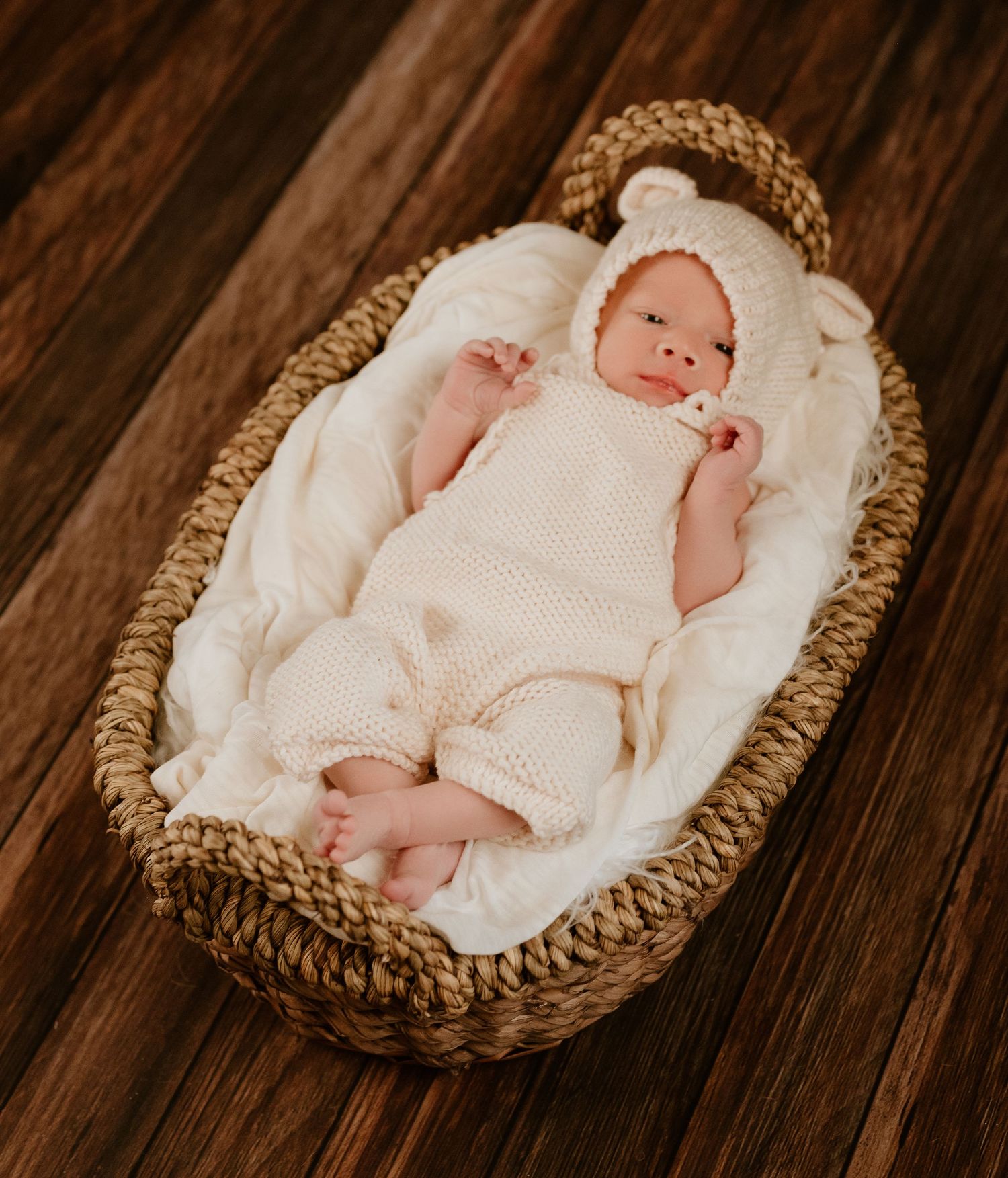 A sleeping newborn wearing white knit clothing rests peacefully in a woven basket lined with soft white fabric.